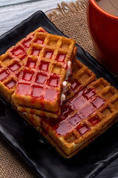 Seitenansicht von Waffeln mit Erdbeersirup auf einer schwarzen Platte und einer Tasse Kakao