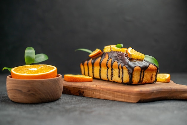 Seitenansicht von weichen Kuchen auf Schneidebrett und geschnittenen Orangen mit Blättern auf dunklem Tisch