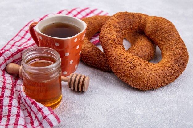 Seitenansicht von weichen türkischen Bagels mit einer Tasse Tee und Honig auf einem Glas auf einem rot karierten Tuch auf einem weißen Hintergrund