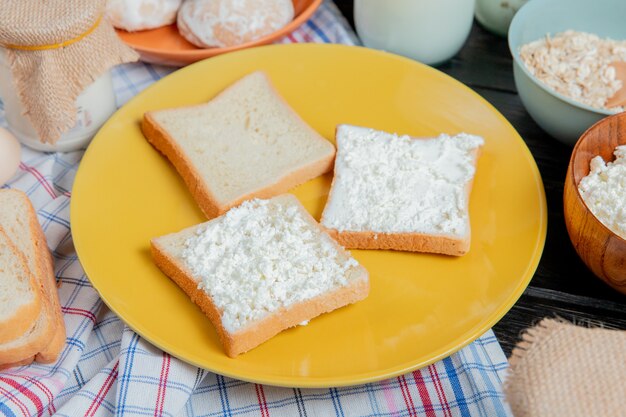 Seitenansicht von Weißbrotscheiben, die mit Hüttenkäse in Teller mit Lebkuchen-Haferflockencreme auf kariertem Stoff und Holzoberfläche verschmiert sind