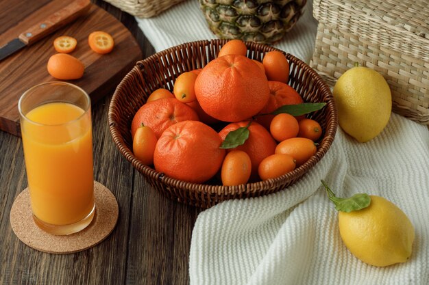 Seitenansicht von Zitrusfrüchten als Mandarine Kumquat in Korbzitronen auf Stoff mit Orangensaft auf Holzhintergrund