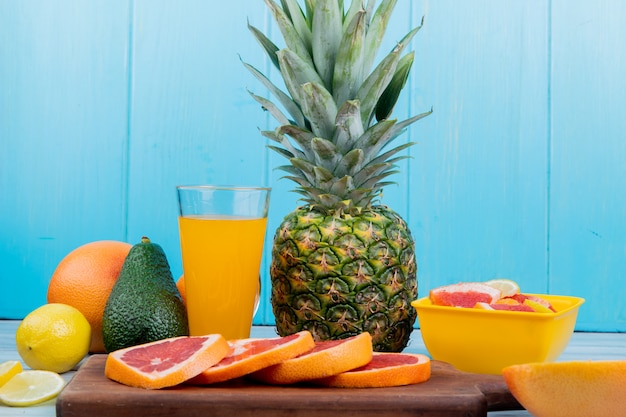 Seitenansicht von Zitrusfrüchten als Zitronen-Avocado-Ananas mit Orangensaft und geschnittener Grapefruit auf Schneidebrett auf Holzoberfläche und blauem Hintergrund