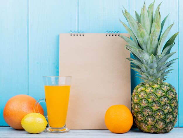 Seitenansicht von Zitrusfrüchten als Zitronengrapefruitorange und -ananas mit Notizblock auf Holzoberfläche und blauem Hintergrund mit Kopienraum