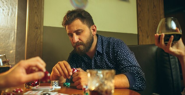 Seitenansichtfoto von männlichen Freunden, die am Holztisch sitzen. Männer spielen Kartenspiel. Hände mit Alkohol Nahaufnahme.