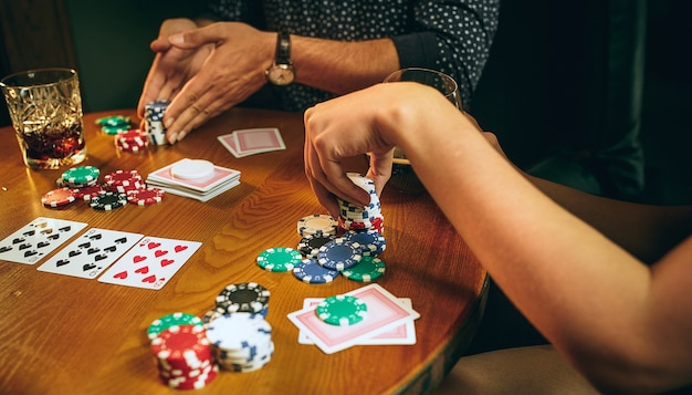 Seitenansichtfoto von männlichen und weiblichen Freunden, die am Holztisch sitzen. Kartenspiel für Männer und Frauen. Hände mit Alkohol Nahaufnahme.