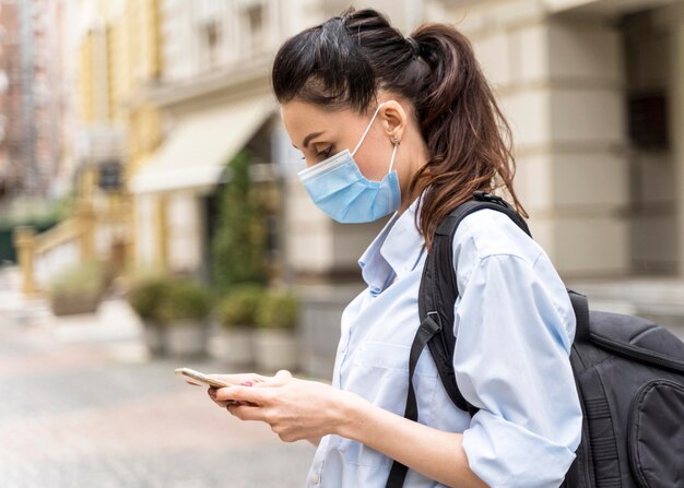 Seitenansichtfrau mit medizinischer Maske, die ihr Telefon prüft