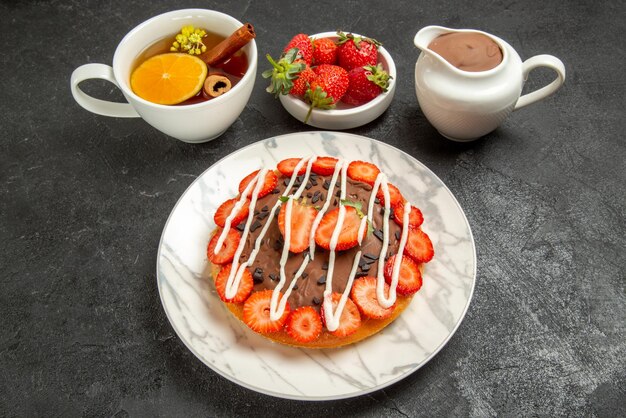 Seitenansichtkuchen mit einer Tasse Teekuchen mit Erdbeeren und Schokolade neben der Schüssel mit Erdbeeren und Schokoladencreme und der Tasse Tee mit Zitronen- und Zimtstangen auf dem schwarzen Tisch