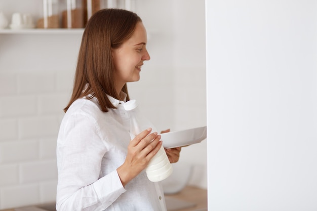 Seitenansichtporträt einer dunkelhaarigen jungen erwachsenen frau, die weißes hemd trägt, lächelnd im kühlschrank schaut, teller in den händen hält, findet morgens eine mahlzeit zum frühstück.