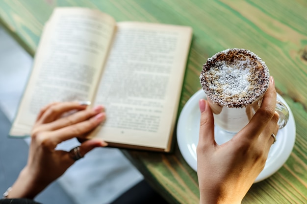 Seitenansichtsfrau, die Latte mit Kokosnuss trinkt und ein Buch am Tisch liest