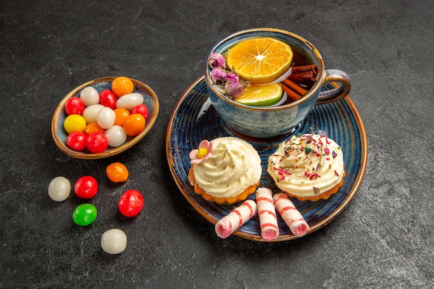 Seitennahaufnahme eine tasse tee blauer teller mit cupcakes mit weißer sahne eine tasse appetitlichen tee und schüsseln mit bunten süßigkeiten auf dem tisch