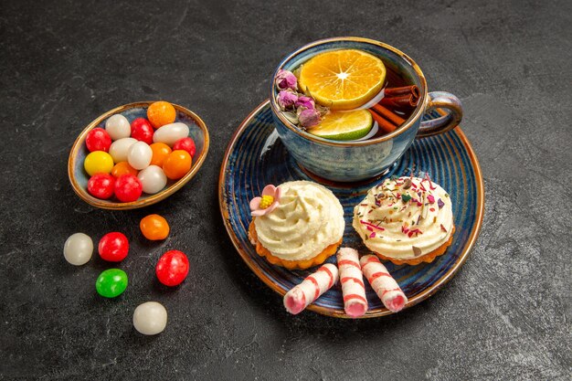 Seitennahaufnahme eine Tasse Tee blauer Teller mit Cupcakes mit weißer Sahne eine Tasse appetitlichen Tee und Schüsseln mit bunten Süßigkeiten auf dem Tisch