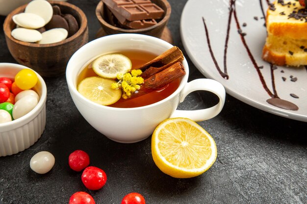 Seitennahaufnahme eine Tasse Tee eine Tasse Tee mit Zitronen- und Zimtstangen Teller mit Kuchen und Schalen mit Schokolade und Süßigkeiten auf dem dunklen Tisch
