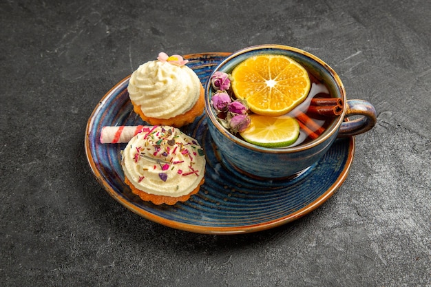 Seitennahaufnahme eine Tasse Tee mit Zitrone zwei Cupcakes mit Sahne und Süßigkeiten neben der Tasse Kräutertee mit Zitrone und Zimt auf dem dunklen Tisch