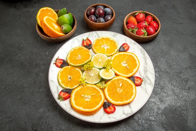 Seitennahaufnahme Früchte auf Tischschalen mit Zitrusfrüchten und Beeren und Teller mit schokoladenüberzogenen Erdbeeren und Zitrusfrüchten in der Mitte des dunklen Tisches