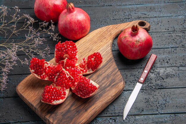 Seitennahaufnahme gepillter Granatapfel und Zweige gepillter Granatapfel auf Küchenbrett neben Baumzweigmesser und drei Granatäpfel auf dunklem Tisch