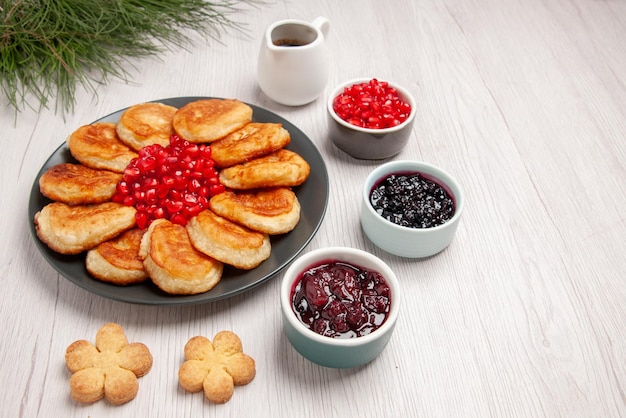 Kostenloses Foto seitennahaufnahme pfannkuchen pfannkuchen und granatapfel auf dem teller neben den schalen mit beeren-keks-sauce und weihnachtsbaum auf dem tisch