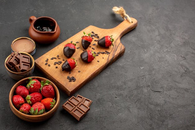 Seitennahaufnahme Schokoladencreme-Erdbeeren und Schokoladencreme und schokoladenüberzogene Erdbeeren auf dem Küchenschneidebrett auf dem dunklen Tisch