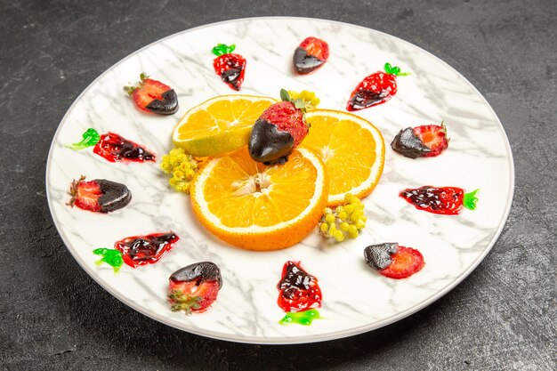 Seitennahaufnahmefrüchte auf dem Teller appetitliche schokoladenüberzogene Erdbeeren und in Scheiben geschnittene Orange auf dem weißen Teller auf dem dunklen Tisch