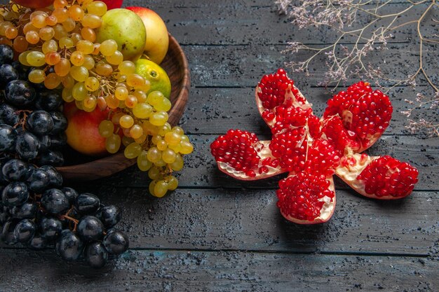 Seitennahaufnahmefrüchte in tellerbrauner Platte mit weißen und schwarzen Trauben Limetten Birnen Äpfel neben Granatapfel und Ästen auf grauem Hintergrund