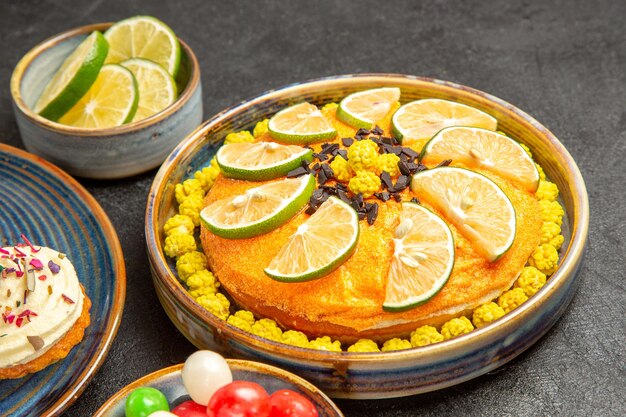 Seitennahaufnahmekuchen mit Limetten ein Teller Kuchen mit Limetten neben der Schüssel mit Süßigkeiten und Limettenscheiben und dem Cupcake auf der blauen Untertasse auf dem Tisch
