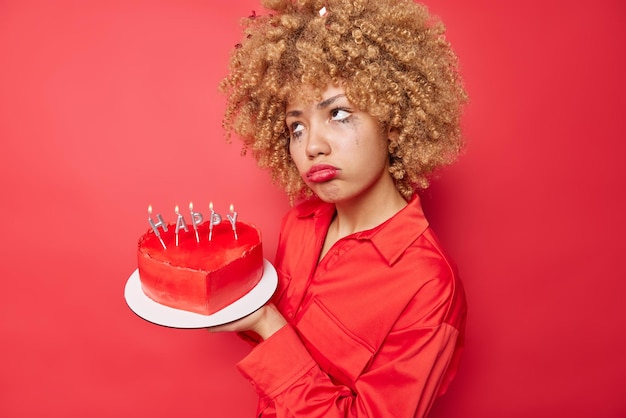 Seitlich erschossen verärgert Frau hat Make-up verwöhnt trägt Kleid hält Herzkuchen mit Kerzen fühlt sich niedergeschlagen isoliert über rotem Hintergrund feiert Valentinstag allein Verwöhntes Urlaubskonzept