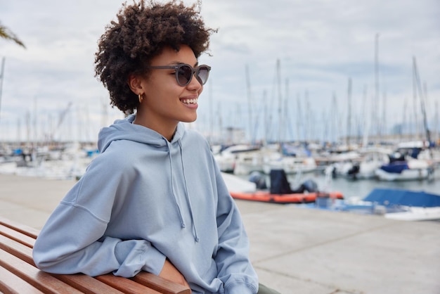 Seitliche Aufnahme einer positiv gelockten Frau, die auf einer Bank posiert, trägt eine Sonnenbrille und ein lässiges blaues Hoodie-Lächeln, das im Großen und Ganzen einen schönen Blick auf den Hafen genießt, atmet frische Meeresluft ein und ruht sich nach dem Gehen aus