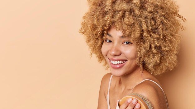 Seitliche Aufnahme einer positiven Frau mit lockigem Haar, die eine trockene Körperbürste verwendet, möchte glatte, saubere Haut haben, lächelt gerne und genießt einen Teil der morgendlichen Gesundheits- und Energieroutine, die vor braunem Hintergrund posiert