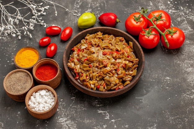 Seitliche nahaufnahme appetitlicher teller grüne bohnen und tomaten in der holzplatte neben den tomaten mit stielen und schalen mit bunten gewürzen auf dem dunklen tisch