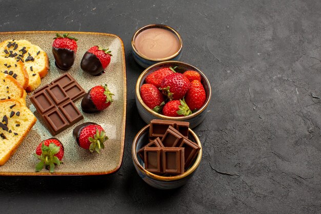 Seitliche Nahaufnahme Dessert appetitlicher Kuchen mit Erdbeeren und Schokolade auf grauem Teller neben Schüsseln mit Erdbeeren und Schokoladencreme auf dunklem Hintergrund