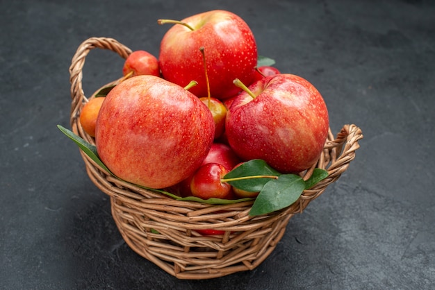 Seitliche Nahaufnahmefruchtfrüchte Äpfel und Kirschen mit Blättern im Korb auf dem dunklen Tisch