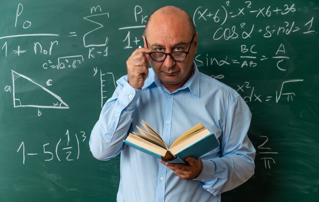 Selbstbewusst aussehende Kamera, männlicher Lehrer mittleren Alters mit Brille, der vor der Tafel steht und ein Buch hält