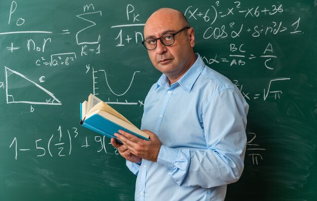 Selbstbewusst aussehende Kamera, männlicher Lehrer mittleren Alters mit Brille, der vor der Tafel steht und ein Buch hält