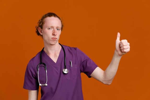 selbstbewusst zeigt Daumen nach oben junger männlicher Arzt in Uniform mit Stethoskop isoliert auf orangefarbenem Hintergrund