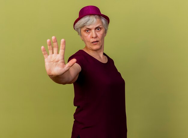 Selbstbewusste ältere Frau, die Partyhutgesten trägt, stoppt Handzeichen lokalisiert auf olivgrüner Wand