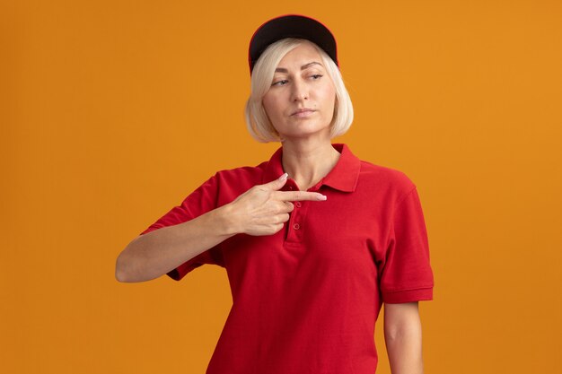 Selbstbewusste blonde Lieferfrau mittleren Alters in roter Uniform und Mütze, die auf die Seite schaut und zeigt