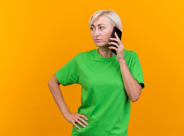 Selbstbewusste blonde slawische Frau mittleren Alters, die am Telefon spricht und die Hand auf der Taille hält, die Seite betrachtet, die auf gelber Wand mit Kopienraum lokalisiert wird