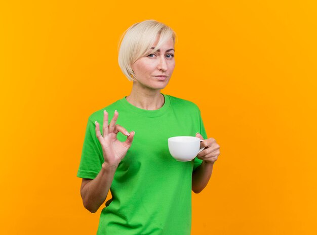 Selbstbewusste blonde slawische Frau mittleren Alters, die eine Tasse Tee hält, die nach vorne schaut, ok Zeichen lokalisiert auf gelber Wand mit Kopienraum