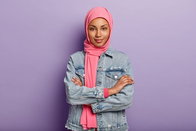 Selbstbewusste dunkelhäutige Frau hat muslimische religiöse Sicht, hält die Arme verschränkt, trägt rosa Kopftuch und Jeansmantel, isoliert über lila Wand, hört Nachrichten mit Interesse