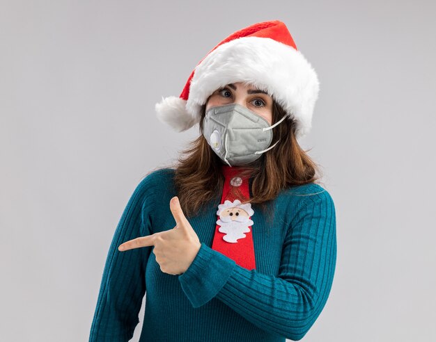 Selbstbewusste erwachsene kaukasische Frau mit Weihnachtsmütze und Weihnachtsmann-Krawatte mit medizinischer Maske, die auf die Seite zeigt, die auf weißer Wand mit Kopienraum isoliert ist isolated