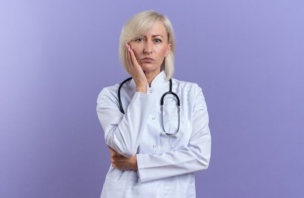 Selbstbewusste erwachsene Ärztin in medizinischer Robe mit Stethoskop, die die Hand auf ihr Gesicht legt und die Vorderseite isoliert auf lila Wand mit Kopienraum betrachtet
