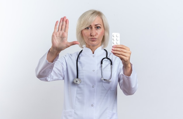 Selbstbewusste erwachsene slawische Ärztin in medizinischer Robe mit Stethoskop, die Stoppschild gestikuliert und Medizintablette in Blisterpackung isoliert auf weißem Hintergrund mit Kopienraum hält