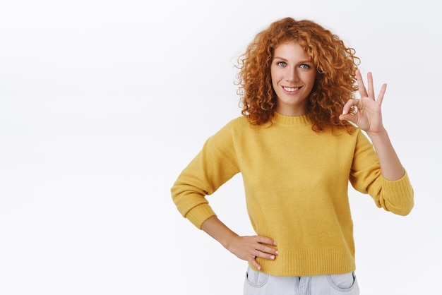 Selbstbewusste, gutaussehende, selbstbewusste, rothaarige, kaukasische Frau mit lockigem Haar, die ein Okay-Zeichen zeigt und mit selbstsicherem Ausdruck lächelt, gibt ihre Zustimmung oder Erlaubnis, die mit einem guten Ergebnis zufrieden ist