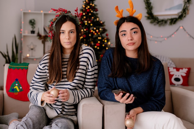 Selbstbewusste hübsche junge Mädchen mit Stechpalmenkranz und Rentierstirnband halten Glaskugelverzierungen, die auf Sesseln sitzen und die Weihnachtszeit zu Hause genießen