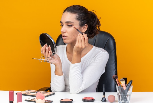 Selbstbewusste hübsche kaukasische Frau, die am Tisch mit Make-up-Tools sitzt und den Spiegel hält und den Lidschatten auf der orangefarbenen Wand mit Kopienraum aufträgt