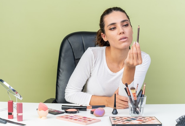 Selbstbewusste hübsche kaukasische Frau, die am Tisch mit Make-up-Tools sitzt und Eyeliner anschaut