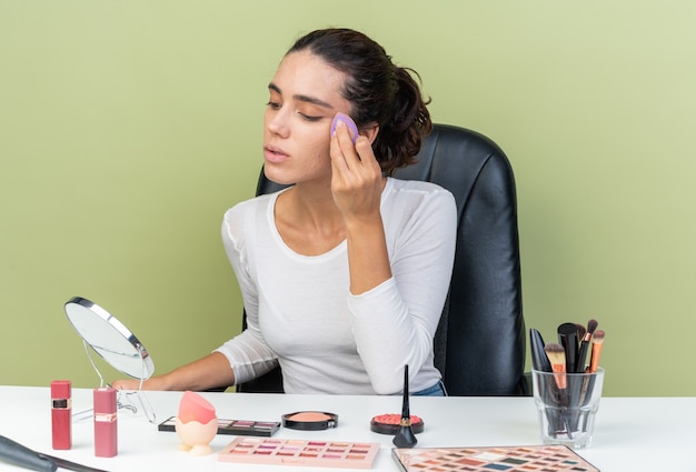 Selbstbewusste hübsche kaukasische Frau, die am Tisch mit Make-up-Tools sitzt und in den Spiegel schaut, der Toncreme mit Schwamm aufträgt