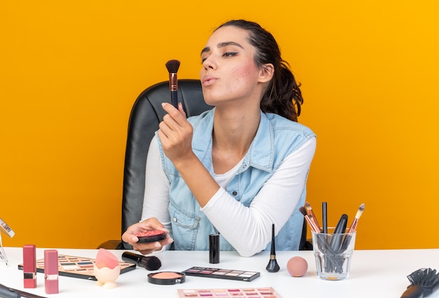 Selbstbewusste hübsche kaukasische Frau, die am Tisch mit Make-up-Tools sitzt und Rouge hält und Make-up-Pinsel betrachtet