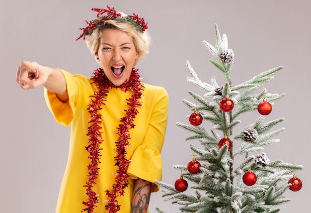 Selbstbewusste junge blonde frau mit weihnachtskopfkranz und lametta-girlande um den hals, die in der nähe des geschmückten weihnachtsbaums steht und gerade schreiend auf weiße wand zeigt