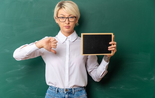 selbstbewusste junge blonde Lehrerin mit Brille im Klassenzimmer, die vor der Tafel steht und eine Mini-Tafel zeigt, die nach vorne schaut und den Daumen nach unten zeigt