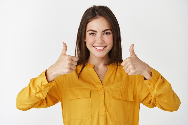 Kostenloses Foto selbstbewusste junge frau, die daumen hoch zeigt und lächelt, positive antwort, ausdrückliche zustimmung, wie und lobt das produkt, das über der weißen wand steht.